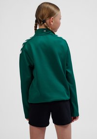 Grøn langærmet skjorte med krave og hvide skulderstriber, lavet af et glat materiale. Kombineret med sorte shorts.