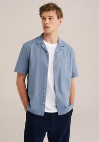 Camisa de manga corta azul claro con cuello, cierre de botones y bolsillo en el pecho. Llevada sobre una camiseta blanca. El fondo es neutral.