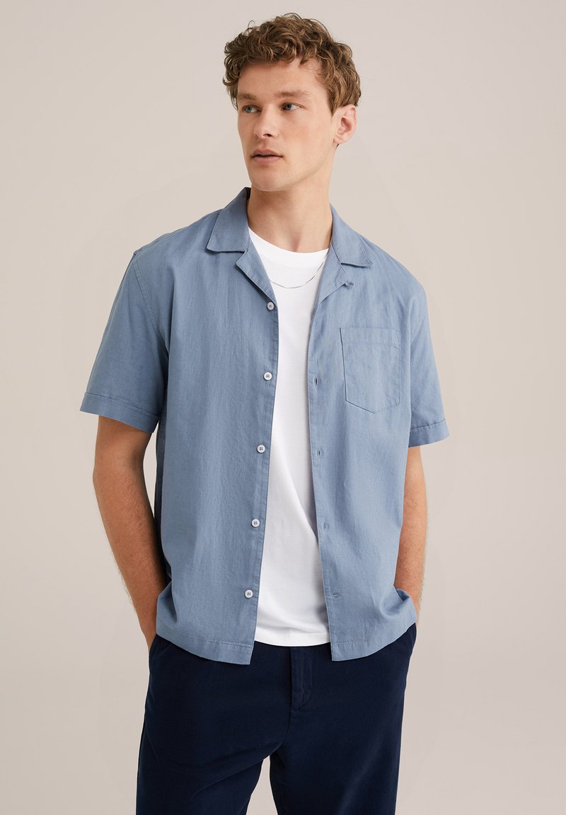 Camisa de manga corta azul claro con cuello, cierre de botones y bolsillo en el pecho. Llevada sobre una camiseta blanca. El fondo es neutral.