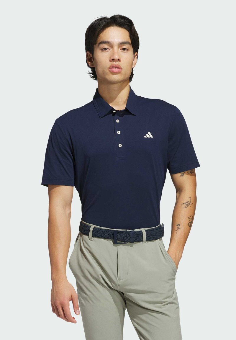Granatowa koszulka polo z kołnierzykiem, z trzema guzikami z przodu i białym logo. Noszona z jasnymi spodniami i plecionym granatowym paskiem.