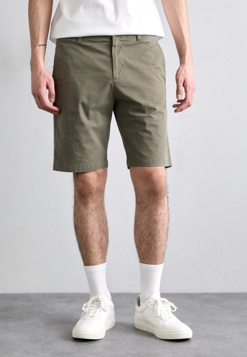 Shorts vert Olive en coton, design ajusté, devant plat, poches latérales, ourlets au genou, associés à des baskets blanches et des chaussettes.