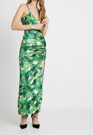 Femme portant une robe maxi à imprimé de feuilles tropicales vertes avec des bretelles fines et une fente latérale, associée à des talons noirs à lanières et des colliers superposés.