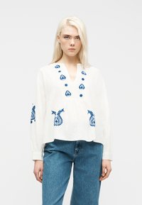 Femme blonde portant un chemisier blanc à manches longues avec des motifs floraux brodés bleus et un jean bleu, debout devant un fond uni.