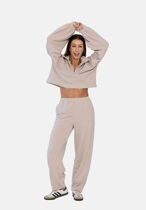 Kobieta uśmiechająca się z podniesionymi rękami, ubrana w beżowy krótki sweter, dopasowane spodnie i białe trampki z czarnymi paskami.