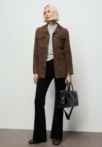 Chaqueta de ante marrón con bolsillos frontales, suéter gris debajo, pantalones acampanados negros y un bolso negro. La modelo posa con un fondo neutro.