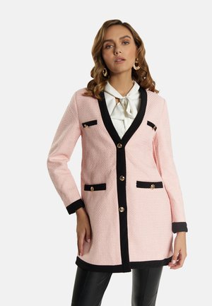 Vrouw draagt een lichtroze textuurjas met zwarte bies en gouden knopen over een witte blouse met een strik, gecombineerd met een zwarte broek.