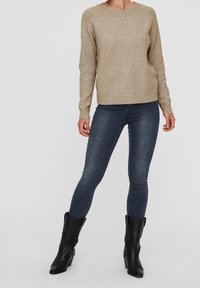 Beiger Strickpullover mit Rundhalsausschnitt und gerippten Bündchen, kombiniert mit dunkelblauen, eng geschnittenen Jeans und schwarzen Leder-Stiefeletten.