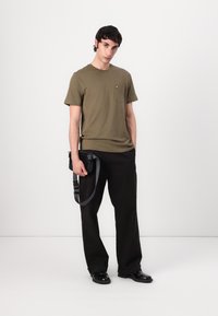 Tricou din bumbac de culoare verde măslinie, cu un buzunar pe piept și un logo brodat galben, asortat cu pantaloni negri și o geantă de umăr neagră.
