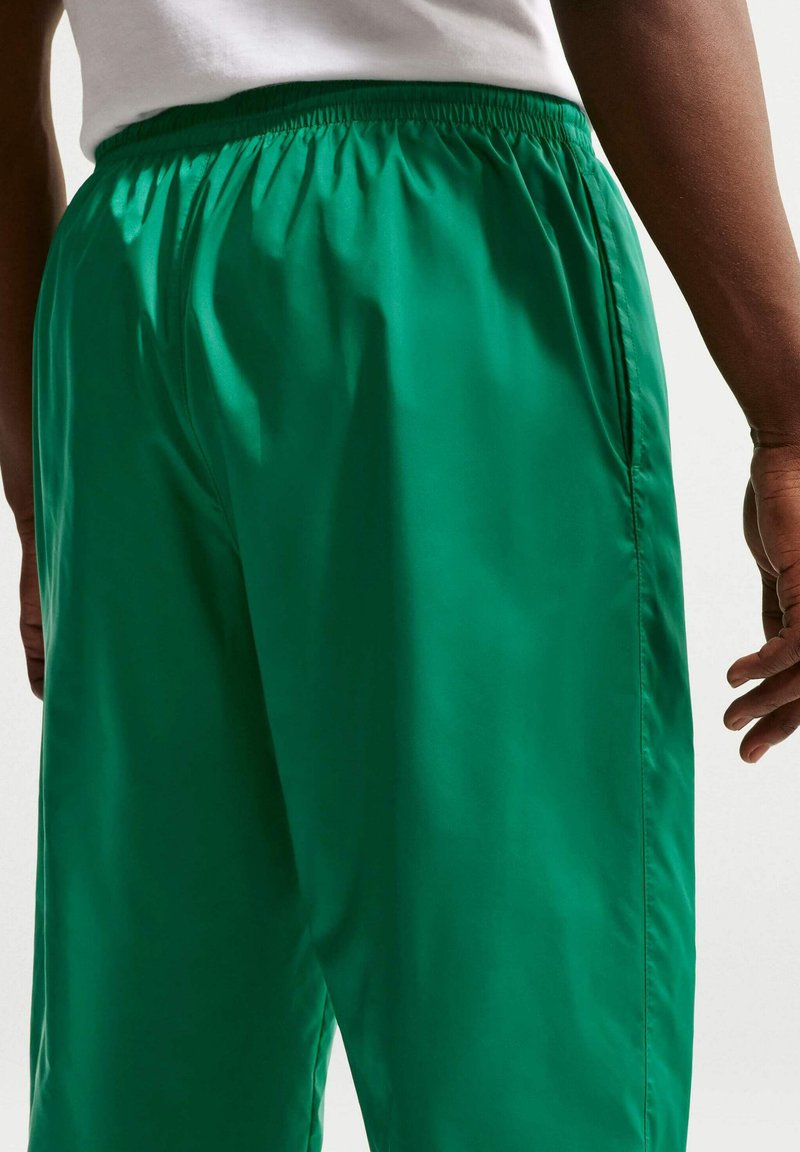 Mann trägt grüne Shorts mit elastischem Bund und weißes Hemd, steht mit entspannten Armen an den Seiten vor einfarbigem Hintergrund.