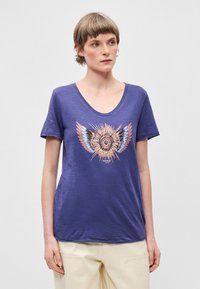 Μπλούζα V με λαιμόκοψη σε μωβ χρώμα, φτιαγμένη από μαλακό ύφασμα, με κεντρικό πολύχρωμο γραφικό σχέδιο που απεικονίζει αφηρημένα φτερά και κυκλικό σχέδιο. Κοντά μανίκια.