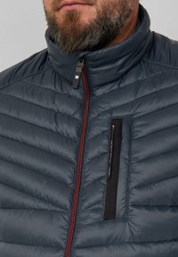 Marineblaue Daunenjacke mit horizontaler Steppung, hohem Kragen und einem Frontreißverschluss mit roten Akzenten. Verfügt über eine schwarze Tasche auf der Brust.