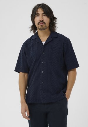Mand med mellemtil langt mørkt hår iført en tekstureret marineblå kortærmet skjorte med knapper og mørke bukser, stående med den ene hånd i lommen.