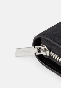 Melnā teksturētā makā ar gludu rāvējslēdzēju, kuram ir sudraba aparatūras birka ar iegravētu "Calvin Klein". Tuvplāns, kas parāda rāvējslēdzēja detaļas.