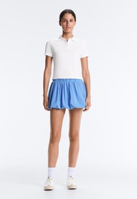 Young woman standing, wearing a white short-sleeve polo shirt, blue striped bubble skirt, white socks, and white sneakers on plain background.