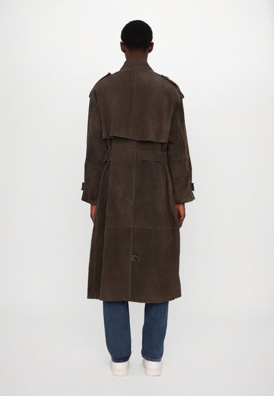 Brauner Wildleder-Trenchcoat mit tailliertem Gürtel, Reverskragen und Schulterklappen; hat ein geschichtetes Rückendesign. Über blauen Jeans getragen.
