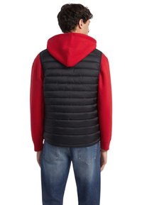 Chaleco acolchado negro con diseño acolchado horizontal, llevado sobre una sudadera con capucha roja. Cuenta con capucha y se combina con jeans azules.