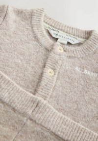 Cárdigan de punto beige con cuello redondo acanalado, cierre de dos botones y logotipo bordado. Textura suave con un patrón sutil.