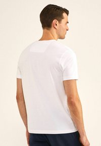 Witte t-shirt met korte mouwen, voorzien van een ronde hals, een gladde textuur en een klein vierkant detail op de achterkant. Gecombineerd met donkere broek.