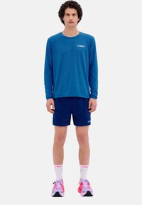 Langärmliges blaues Sportshirt und marineblaue Shorts, mit weißen Logo-Akzenten, ergänzt durch rosa-weiße Laufschuhe und Knö socks.