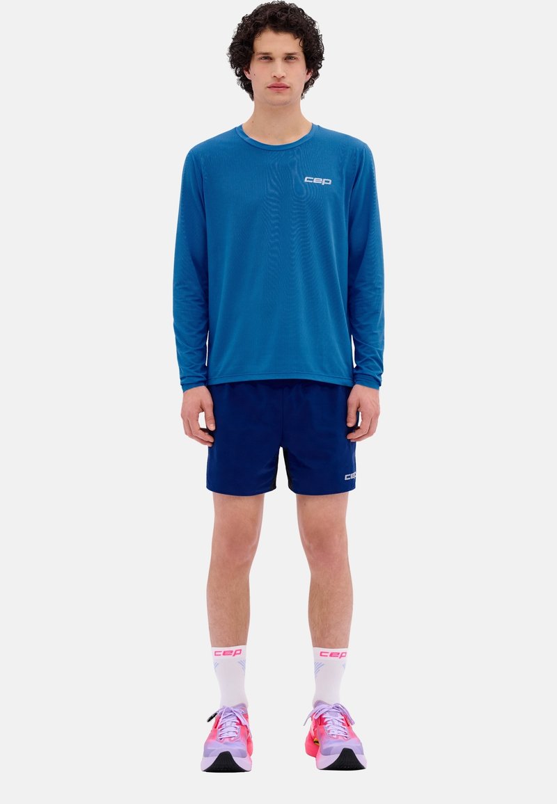 Langärmliges blaues Sportshirt und marineblaue Shorts, mit weißen Logo-Akzenten, ergänzt durch rosa-weiße Laufschuhe und Knö socks.