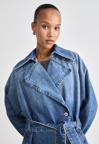Denim trenchcoat in lichtblauw met een grote kraag, een dubbelzijdige sluiting aan de voorkant, een ceintuur in de taille en opvallende stikseldetails.