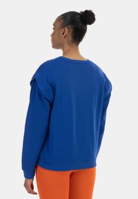 Kék, laza szabású pulóver, bordázott mandzsettákkal és kerek nyakkivágással. A hátoldal sima textúrájú; élénk narancssárga leggings-szel viselve.