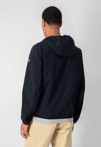 Marinblå huva jacka tillverkad av slät tyg, med elastiska ärmmuddar och en logotyp på vänster ärm. Lätt design, avslappnad passform.