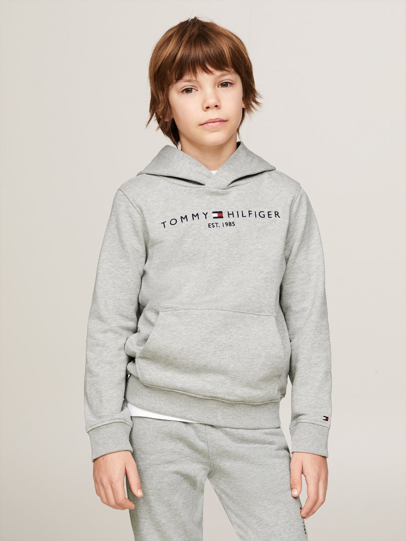 Felpa grigia con cappuccio, coulisse e tasca frontale, con la scritta "TOMMY HILFIGER EST. 1985" in blu scuro. Indossata con pantaloni grigi abbinati.
