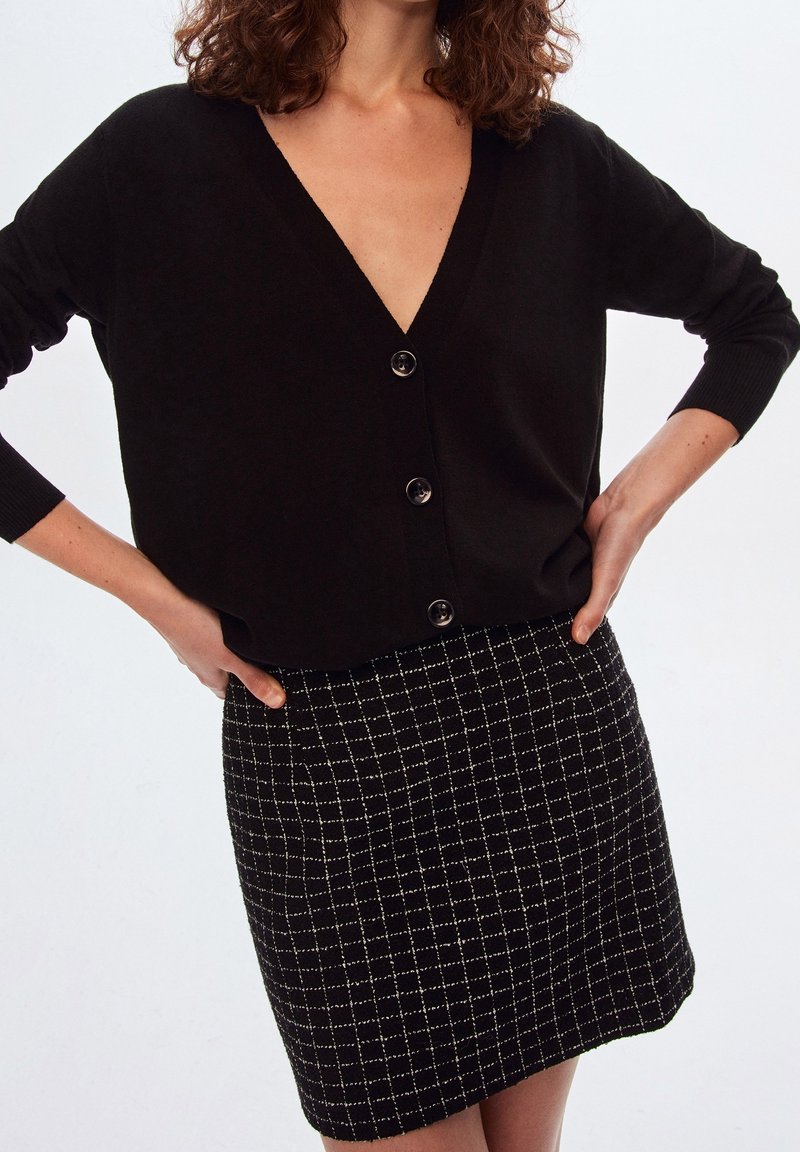 Cardigan noir à col en V avec cinq boutons, associé à une mini-jupe à carreaux noir et blanc avec un motif texturé et une silhouette ajustée.