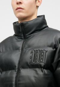 Close-up de uma pessoa a usar uma jaqueta de couro preta acolchoada com um colarinho alto e frente com fecho zip, apresentando um logótipos gótico em forma de "W" costurado.