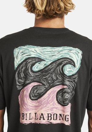 Blonde persoon in een zwart Billabong t-shirt met een abstracte pastelgekleurde golfontwerp op de achterkant.