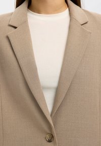 Beige blazer featuring a textured fabric, notched lapels, and a single button closure, layered over a white top.