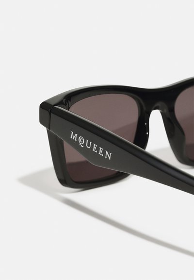Occhiali da sole neri con lenti scure, design della montatura angolare e logo "M⍐QUEEN" sulla tempia. Finitura opaca, aspetto elegante e moderno.