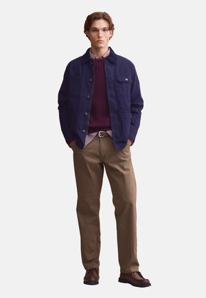Young man stands with hands in pockets wearing navy jacket, burgundy sweater over checked shirt, brown pants, and dark brown shoes on white background.