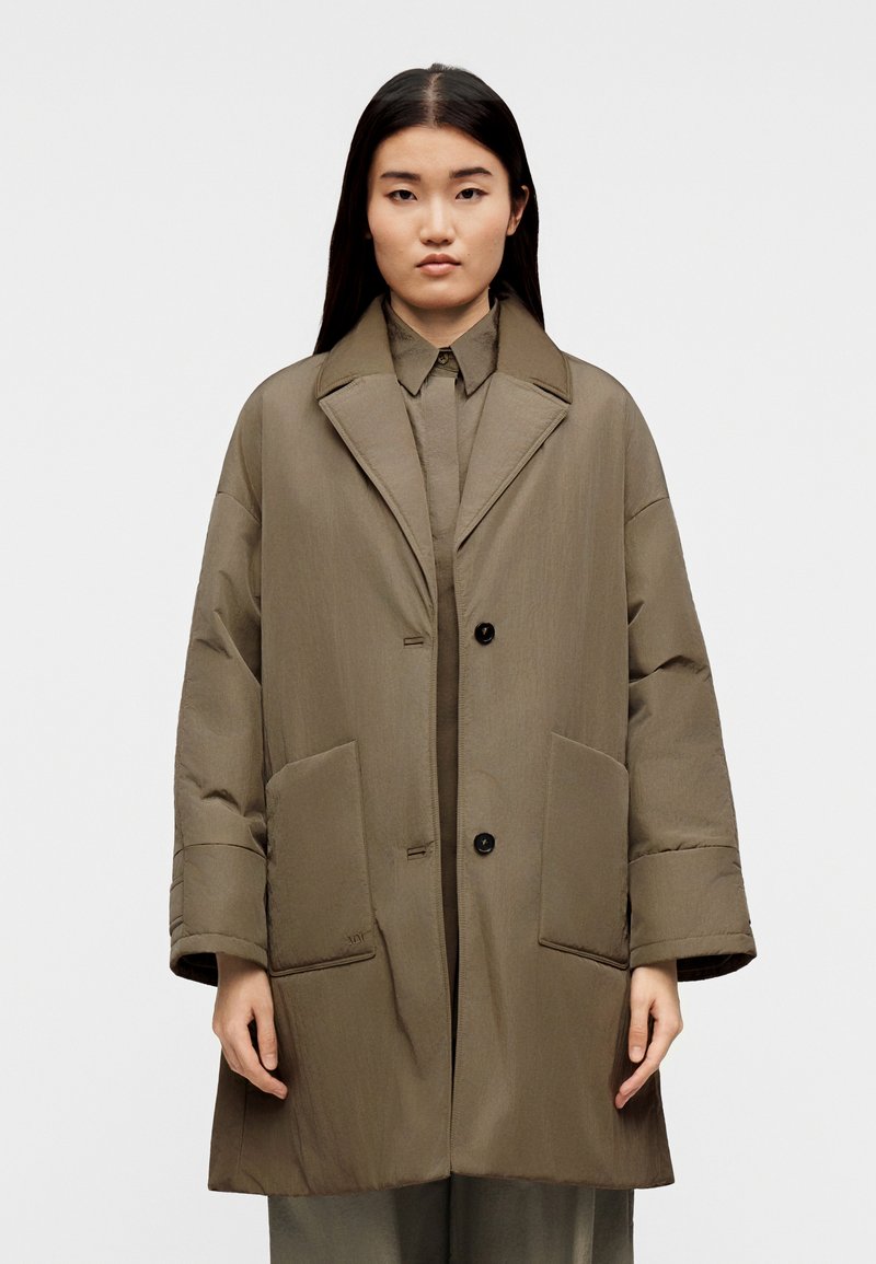 Nő hosszú fekete hajjal, nagy méretű, olívazöld steppelt kabátot visel, amelynek nagyméretű hajtókái és elöl felvarrt zsebei vannak, fehér háttér előtt áll.