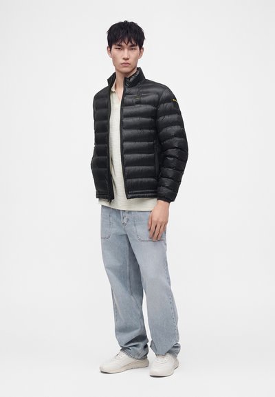 Jeune homme portant une doudoune noire, une chemise beige clair, un jean bleu clair ample et des baskets blanches, debout devant un fond uni.