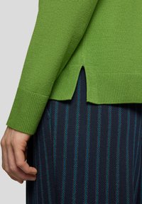 Grüner gestrickter Pullover mit gerippten Bündchen und seitlichem Schlitz. Über einem dunkel gestreiften Stoff mit blauen Akzenten getragen. Nahaufnahme.