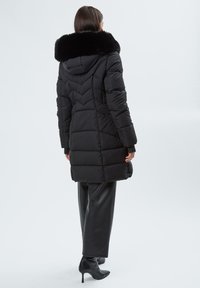 Abrigo acolchado negro con cuello de piel sintética, costuras en forma de chevron, puños acanalados y un diseño de longitud media, combinado con pantalones de cuero y botas de tacón.