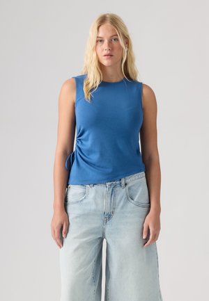Ung kvinde med langt blondt hår iført en ærmeløs blå top og lyseblå jeans med vide ben, stående mod en ensfarvet baggrund.