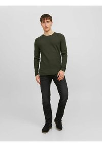 Langærmet olivengrøn strikskjorte med et tekstureret mønster, parret med mørke jeans og sorte sko, stående foran en neutral baggrund.