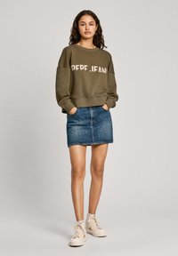 Sudadera verde oliva con grandes letras beige, combinada con una falda de mezclilla azul y zapatillas altas color crema. Ajuste casual y relajado.