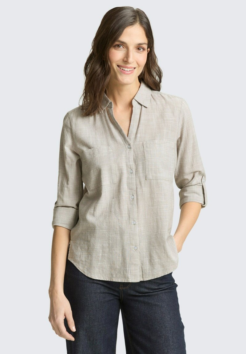 Õlgadeni ulatuvate pruunide juustega naine kannab beežikat nööbitavat särki kergete rullitud varrukatega ja tumedat sinist teksaseid, naeratab ühe käega taskus.