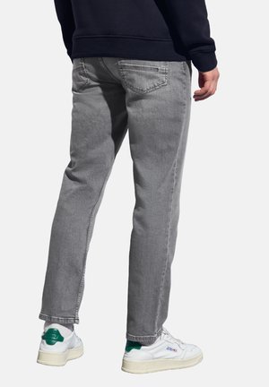 Persoon in grijze jeans, witte sneakers met groene accenten en een donkere sweater, gezien vanaf de achterkant tegen een witte achtergrond.