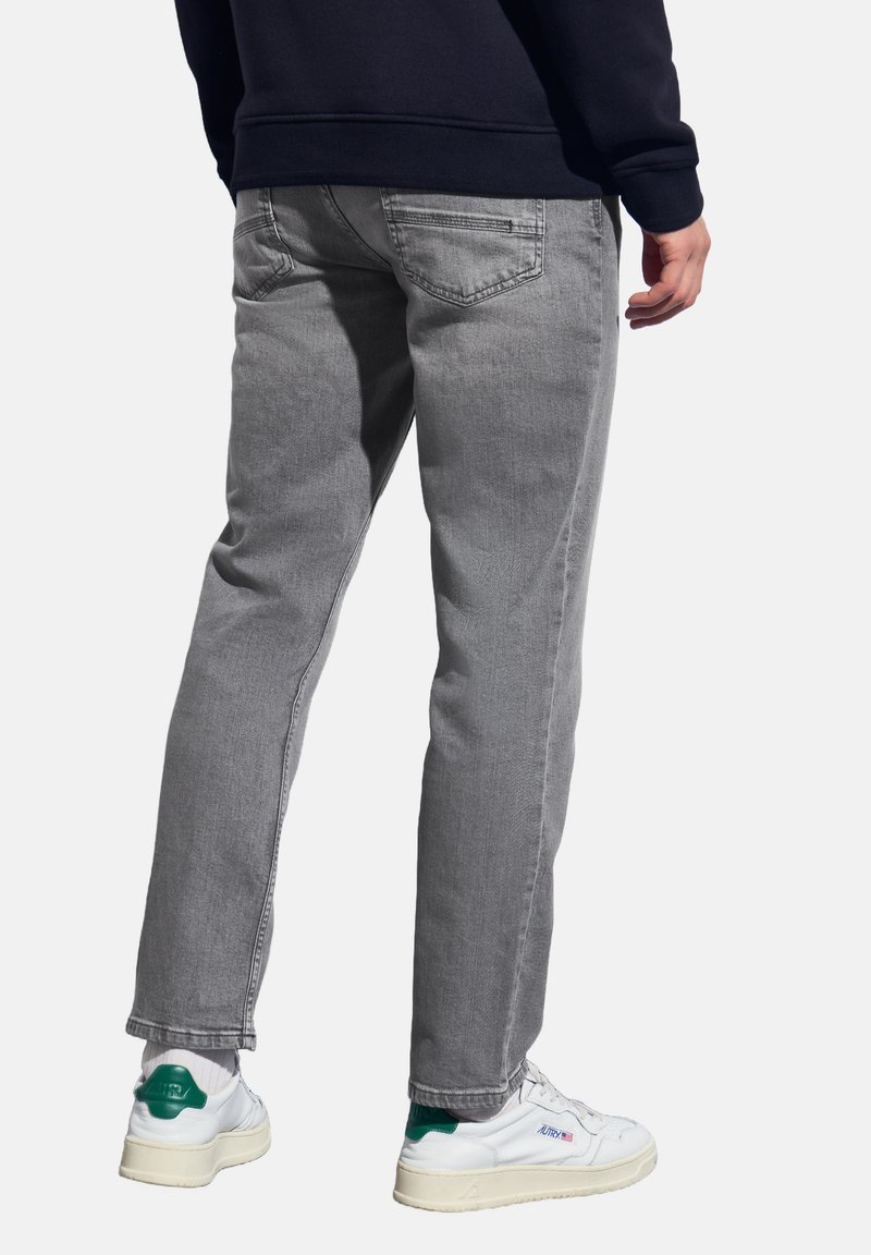 Persoon in grijze jeans, witte sneakers met groene accenten en een donkere sweater, gezien vanaf de achterkant tegen een witte achtergrond.