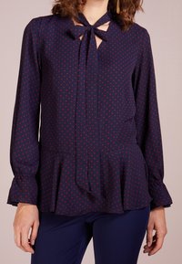 Marinblus med röda prickar, långa ärmar, knytkrage och peplumslut. Slätt tyg med en lätt lyster och instoppade ärmslut.