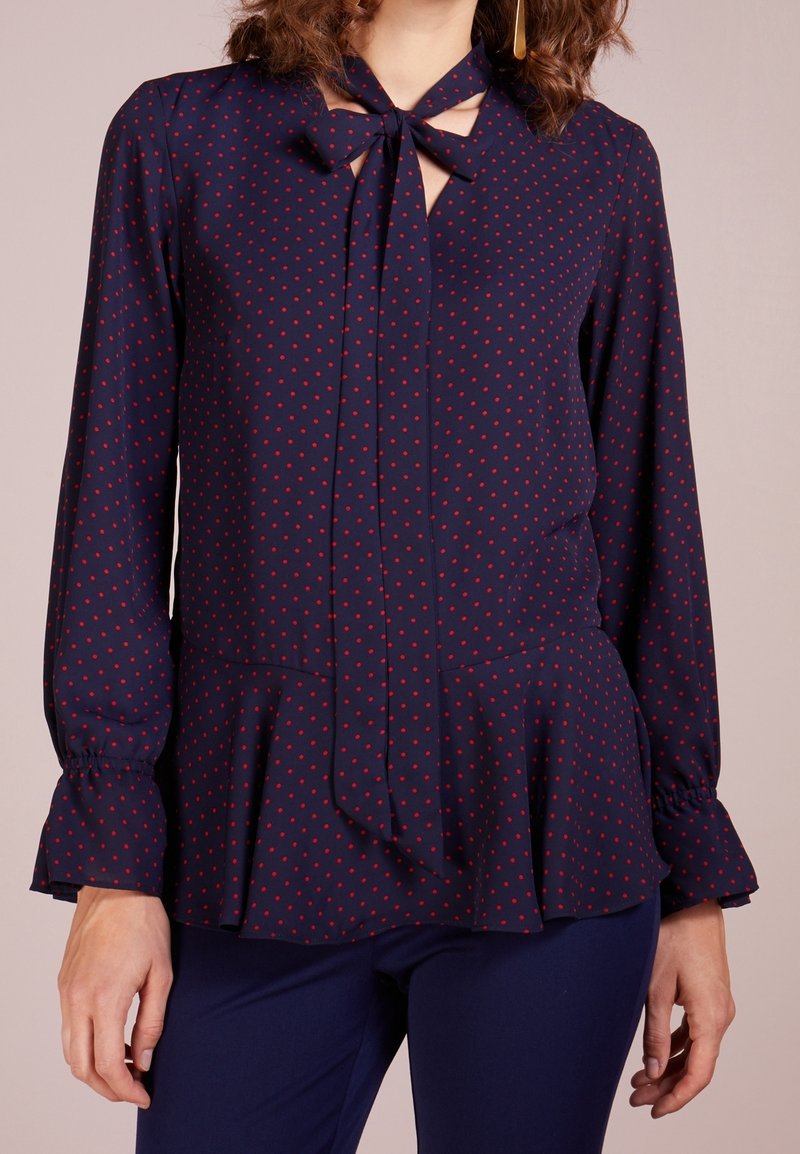 Marinblus med röda prickar, långa ärmar, knytkrage och peplumslut. Slätt tyg med en lätt lyster och instoppade ärmslut.