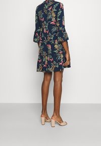 Blommig klänning med marinblå bakgrund, volangärmar och lagerdesign. Har rosa och gröna mönster. Bärs med nakna blockklack-sandaler.