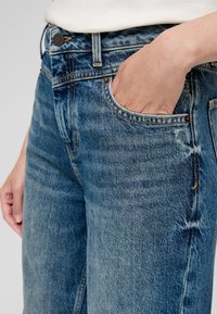 Niebieskie dżinsy mają luźny krój, wypłowiały teksturę i widoczne szwy. Krawędź przedniej kieszeni jest zaokrąglona; obecny jest mosiężny guzik.