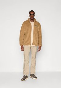 Barna bolyhos dzseki cipzárral, alatta bézs póló, bézs cargo nadrág, valamint szürke, sárga részletekkel díszített sportcipő. Napszemüveggel kiegészítve.