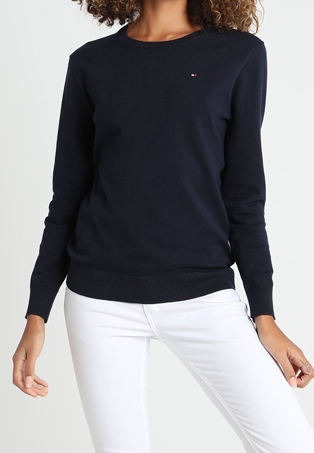 Kvinna klädd i en skräddarsydd marinblå långärmad tröja och vita smala byxor, står med en hand avslappnad och den andra något upphöjd.